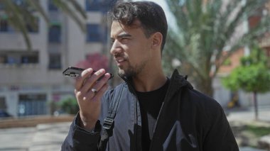 Parkta palmiye ağaçları ve binaları olan bir telefona konuşan genç bir İspanyol açık hava kentsel ortamını sergiliyor..