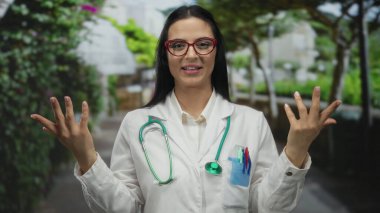 Steteskoplu İspanyol kadın doktor açık hava ortamında parlak, yapraklı bir park ortamında gülümsüyor, sağlık ve sağlık hizmeti sunuyor..