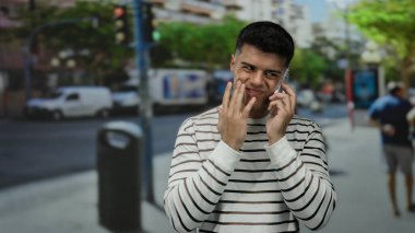 Kalabalık bir şehir caddesinde telefon konuşması yapan genç adam, arka planda şehir araçları ve yayalar ile canlı ifadeler sergiliyor..