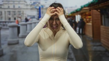 Stresli bir kadın, açık hava pazarında, kafası hayal kırıklığıyla dolu, yoğun bir alanda bej bluz giyiyor ve bulutlu bir gökyüzünün altında arka planı bulanık..