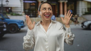 Middle aged woman in white shirt with hands raised and broad smile on busy city street; happy surprise.