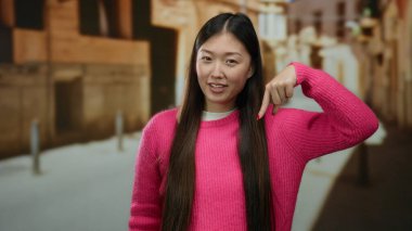 Canlı pembe kazak giyen bir kadın arka planda bulanık binalar olan bir şehir caddesini işaret ederek neşeli bir ifade sergiliyor..