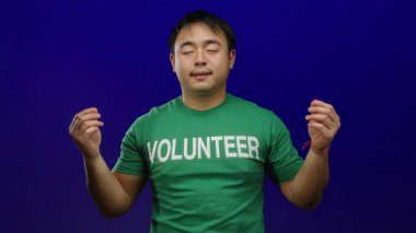 Genç bir adam yeşil bir gönüllü gömleği içinde, canlı mavi bir arka plana karşı huzur içinde meditasyon yapıyor, sakinlik ve odaklanma hissi uyandırıyor..