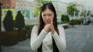 Sokak sokak gezen, ciddi jestleri olan, Asyalı kökenli, uzun saçlı, şehir manzarasında beyaz bluz giyen bir kadın..