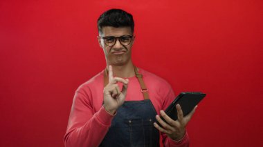Gözlük takan ve önlük takan genç adam tabletle canlı kırmızı arka planda etkileşime giriyor. Modern teknoloji ve gündelik stil göz alıcı bir şekilde izole edilmiş..