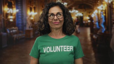 Müze salonunda orta yaşlı İspanyol kadın gönüllü yeşil tişört giyip dil uzatıyor, mizah nişanı yapıyor..