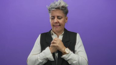 Mor stüdyoda elini göğsüne koyan kısa gümüş saçlı bir kadın ciddi bir ifadeyle; sessiz endişe yansıması.