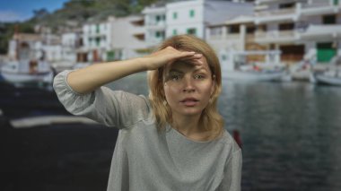 Gözleri gölgeleyen kadın ve güneşli mavi sahil limanı sahnesinde gülümseyen kadın; beklenti.