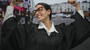 Dışarıda yargıç kılığında gülümseyen bir kadın şehrin arka planında coşkulu bir jest yaparak kollarını kaldırıyor. Kendine güveni ve başarısı tam..