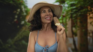 Bikinili İspanyol kadın yeşil bir parkta dondurmadan hoşlanıyor, güneş şapkası takıyor ve canlı bir yaz ortamında mutlu bir şekilde gülümsüyor..