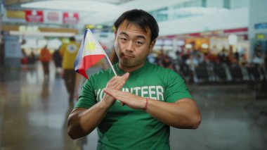 Yeşil gönüllü gömlekli genç adam havaalanı terminalinde filippine bayrağı taşıyor, elleriyle el kol hareketi yapıyor, kültürel çeşitliliği somutlaştırıyor ve içeride gönüllü ruh oluşturuyor..