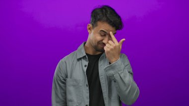 İzole edilmiş mor arka planda gözlerini ovuşturan genç bir adam yorgunluk ve stresi çağrıştırıyor, şehir profesyonel ortamında duyguları ve ifadeleri vurguluyor.