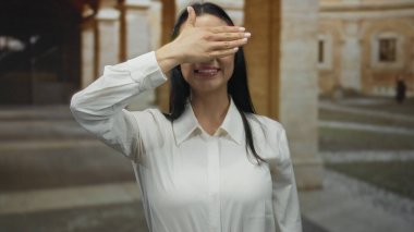 Eski bir üniversite avlusunda gülümserken gözlerini kapayan genç bir kadın mimari arka planda neşeli bir an yaşatıyor..
