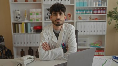 Tıbbi malzeme ve dosyalarla çevrili bir klinikteki bir masada oturan laboratuvar önlüğü giyen genç adam profesyonellik ve özen gösteriyor..