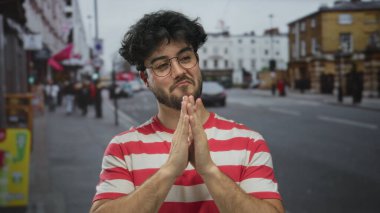 Çizgili tişörtlü, sakallı, genç İspanyol bir adam kalabalık bir şehir sokağında elleri birarada duruyor. Düşüncelerini ya da minnettarlığını doğal ışıkla ifade ediyor..