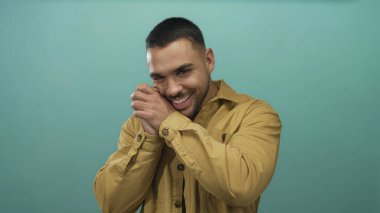 Hardal tişörtü giyen İspanyol genç adam gülümser ve ellerinin yanaklarını deniz mavisi stüdyo ortamında bastırır; sıcaklık mutluluğu.