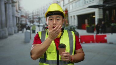 İnşaat işçisi, elinde sert şapka ve güvenlik yeleği olan bir kahveyle şehir sokaklarında esniyor..