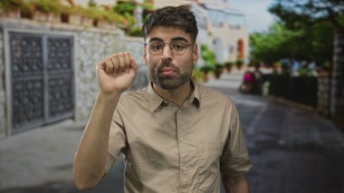 Taştan duvarlar ve yeşil ağaçlarla kaplı güneşli bir caddede omuz silkme hareketi yapan bir adam..