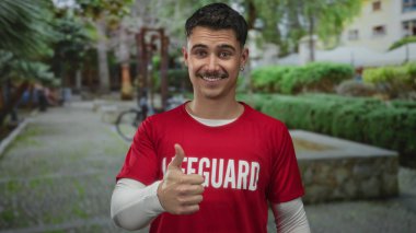Kırmızı cankurtaran tişörtlü genç adam gün boyunca canlı bir açık hava parkında başparmağıyla gülümsüyor..