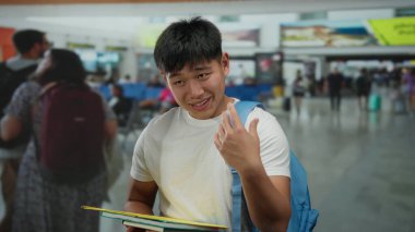 Elinde seyahat belgeleri olan genç Çinli bir adam hareketli bir havaalanı terminalinde duruyor, beklenti ve küresel keşif duygusunu iletiyor..