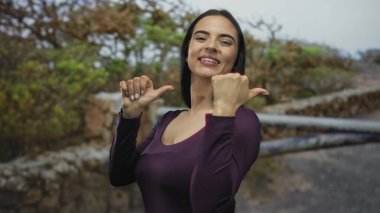 Parktaki kadın başparmaklarını yemyeşil bir gülümseme ile geriye doğru gösteriyor bulutlu bir gökyüzünün altında mor bir gömlek giyiyor..