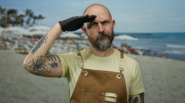 Güneşli bir günde kumlu sahilde dalgalar arkadan vururken kahverengi tulumlu, sakallı ve dövmeli bir adam selamlıyor.