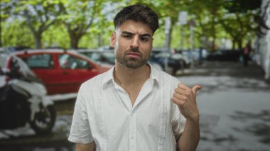 Adam, park halindeki arabalar ve yapraklı ağaçlarla dolu işlek bir caddede başparmağını omzunun üzerinden uzatıyor..