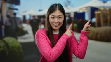 Pembe kazaklı bir kadın kalabalık bir pazar yerini işaret ediyor, insanlarla çevrili ve açık hava tezgahları, canlı alışveriş deneyimi öneriyor..