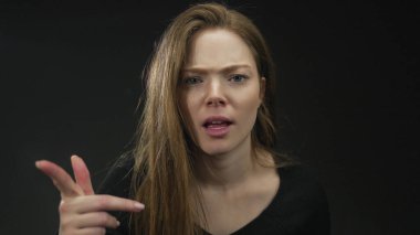 Siyah stüdyo ortamını gösteren uzun kahverengi saçlı, suratsız, beyaz bir kadın..