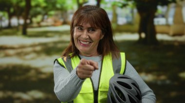 Latin kökenli son sınıf öğrencisi bir kadın parkta kameraya bakarken, yansıtıcı bir yelek giyip, bisiklet kaskı tutarken, açık hava aktivitelerinde güvenliği somutlaştırırken gülümsüyor..