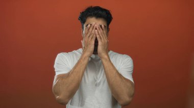 Koyu renk saçlı, sakallı ve beyaz gömlekli kaşları çatık bir adam turuncu stüdyoda boynu yukarı doğru eğilmiş..