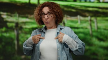 Gözlüklü bir kadın yeşil bir parkta dikilirken şaşkınlıkla kendini işaret ediyor..