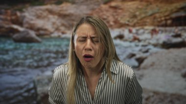Woman grips lower back and grimaces with eyes closed against beach backdrop in studio standing; pain.