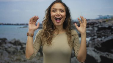 Heyecanlı yüzlü bir kadın bulutlu bir gökyüzünün altında kayalık bir sahil kenarında duruyor. Elleriyle doğanın güzelliğini ve duygularını gösteriyor..