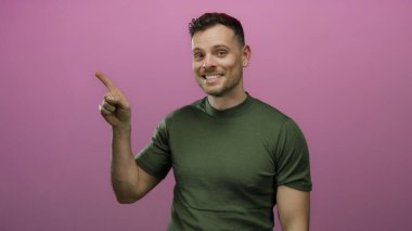 Gülümseyen ve coşkuyla işaret eden genç bir adam canlı pembe bir arka planda izole edilmiş bir stüdyoda neşeli ifadeler ve kendinden emin tavırları ortaya koyuyor..