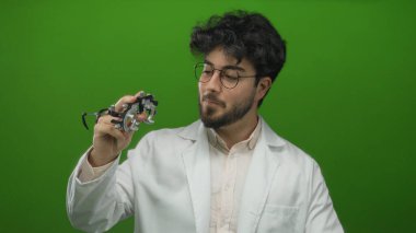 Sakallı ve gözlüklü genç adam canlı yeşil bir arkaplan karşısında optometri ekipmanı tutuyor, beyaz önlük giyiyor, stüdyo ortamında kendine güvenen bir gülümseme sergiliyor..