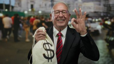 Takım elbiseli kıdemli adam elinde dolar sembolü olan para çantası tutuyor ve kalabalık bir sokakta iyi bir jest yapıyor..