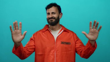 Turuncu hapishane tulumu giyen İspanyol sakallı adam mavi arka planda duruyor. Elleri havada ve sakin bir şekilde teslim ol ya da dur senaryosu öneriyor..