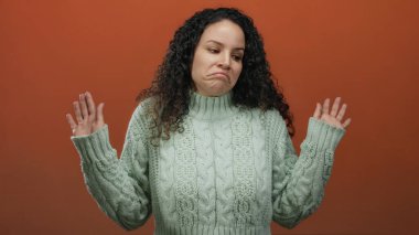 Yeşil kazaklı, kıvırcık saçlı bir kadın, izole edilmiş turuncu bir arka plana karşı kayıtsızlığını dile getiriyor. Stüdyo ortamında omuz silkme hareketi yapıyor..