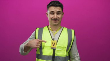 Yansıtıcı yelek giyen genç İspanyol adam, izole edilmiş pembe duvar arka planına karşı gönüllü rozeti işaret ediyor.