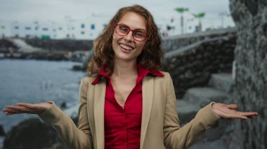 Sahilde okyanus manzaralı, kayalık manzaralı, kırmızı bluz ve bej ceketli bir kadın sahilde neşe ve rahatlık gösteriyordu..