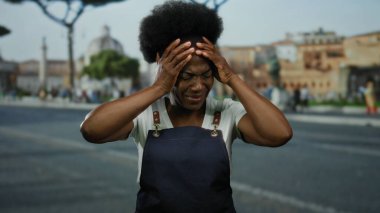 Önlüklü bir kadın şehir caddesinde hayal kırıklığı içinde başını tutuyor. Bulanık binalar ve ağaçlarla şehir zeminine karşı stresi dile getiriyor..