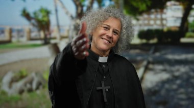 Şehir ortamında gri saçlı, sıcacık gülümseyen yaşlı bir kadın, önemli bir haç kolyesiyle papaz kıyafeti giyiyor, elini davetkar bir şekilde uzatıyor..