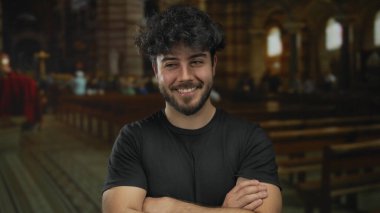 Sıradan giysiler içinde sakallı genç İspanyol bir adam kollarını kavuşturup duruyor loş aydınlatılmış bir kilisede ahşap sıralar ve vitray pencereler var..