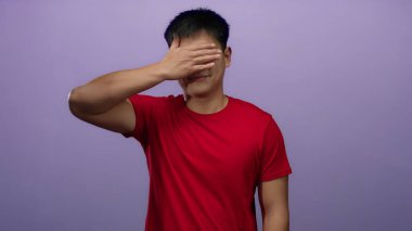 İzole edilmiş mor arka planı olan yüzü kaplayan genç bir adam kırmızı bir gömlek giyiyor, neşeli ve canlı bir ifade sergiliyor..