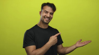 Siyah tişörtlü genç İspanyol bir adam avucunun içini göstererek izole edilmiş sarı bir arkaplanı işaret ediyor. Oyuncu bir jest olarak sunum veya reklam önerisinde bulunuyor.