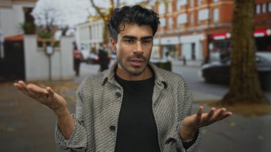 Erkek, çatık kaşlarıyla omuz silkerken bir sokak ortamında göğsünün önünde ellerini açık tutar; hayal kırıklığı.