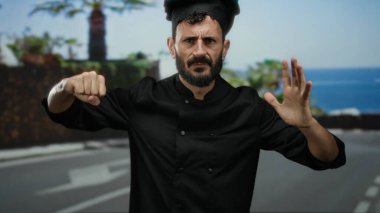 Siyah üniformalı İspanyol şef, güneşli bir sokakta yumruklarıyla enerji dolu jestler yapıyor. Palmiye ağaçları ve mavi gökyüzü arkaplanı arasında kararlılık ve duyguyu gösteriyor..