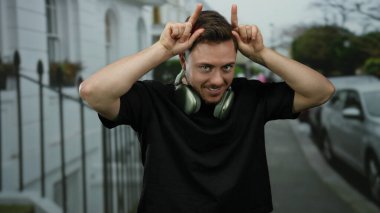 Sakallı ve kulaklıklı genç adam arka planda bulutlu gökyüzü ile şehir caddesinde neşeli el hareketleri yapıyor..