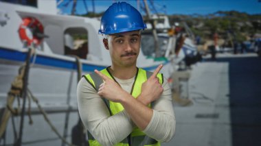 Yansıtıcı yelek giyen ve kasketli genç bir adam arka planda bir tekneyle kıyıya bakan bir limanda duruyor..